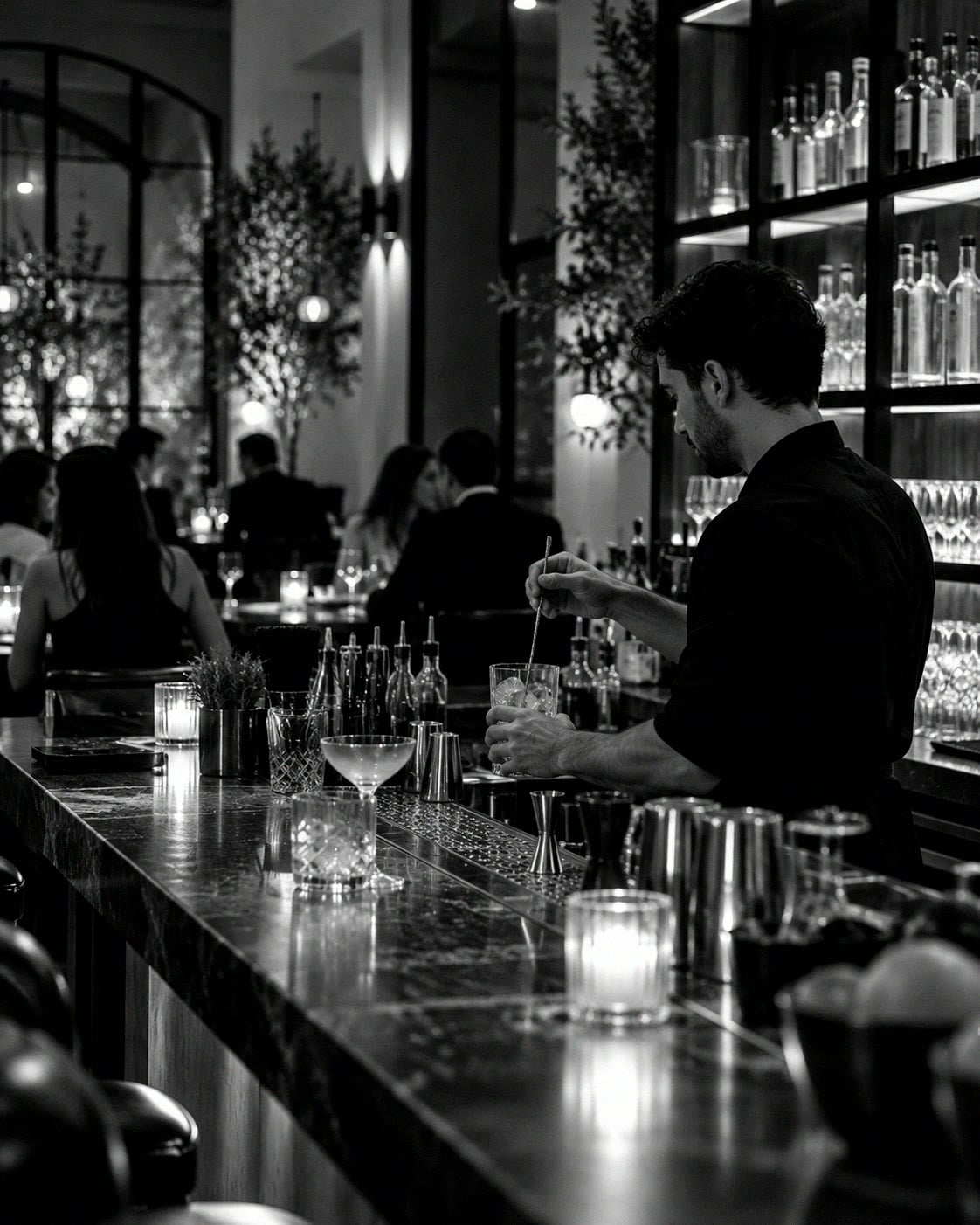 A bartender stirring a drink at a cocktail bar early in the evening, shelves of glasses behind.
