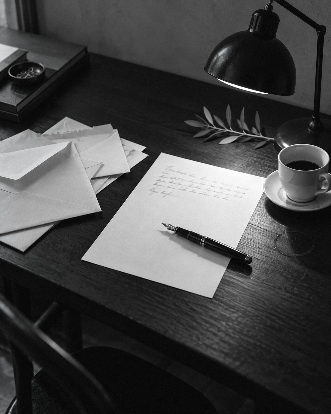 A reply half-written with a fountain pen on a dark desk, beside opened letters, a lamp and a cup of coffee.